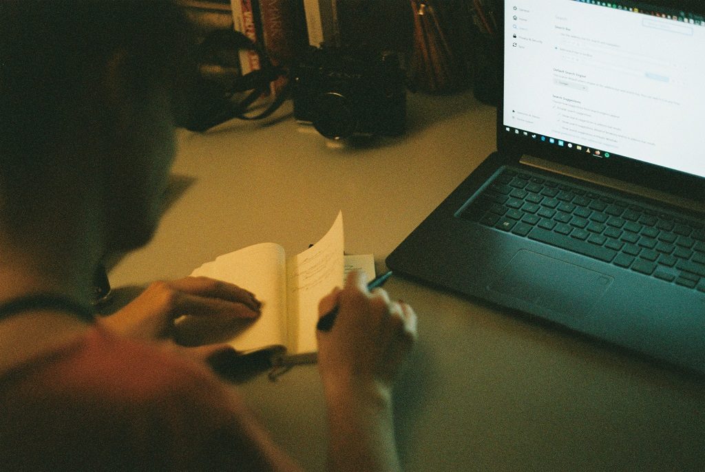 Student makes some notes in a pocketbook from a screen of a laptop 