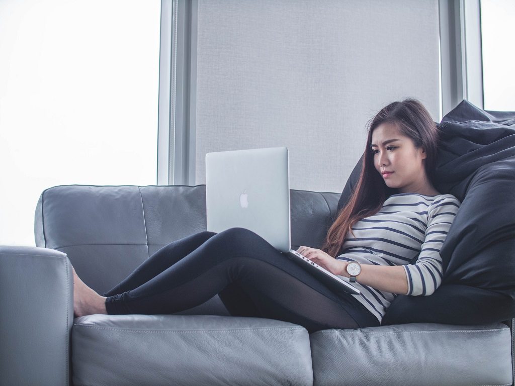 Student girl sitting on sofa and working remotely with laptop on her knees.